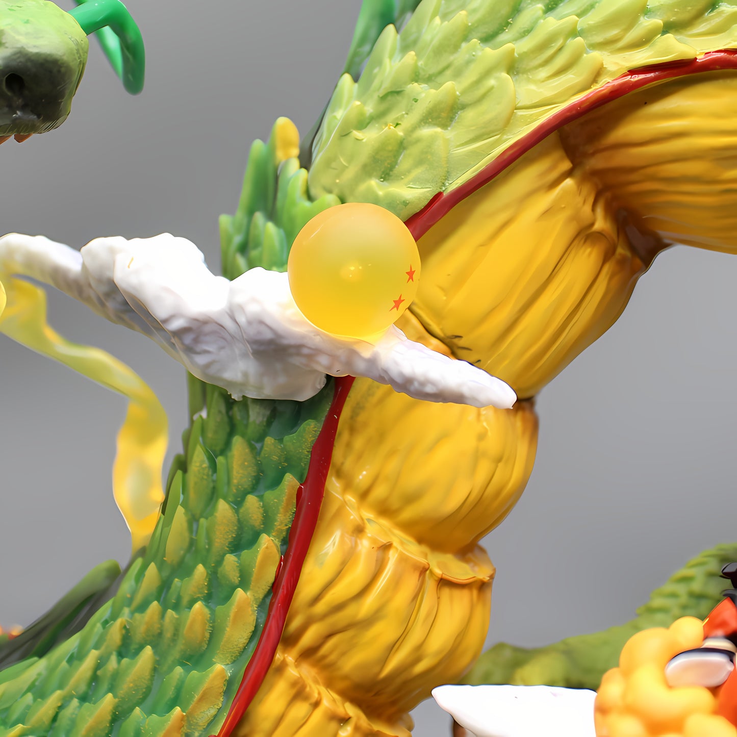 A close-up of a segment of the Shenron dragon figurine from Dragon Ball Z, showcasing a translucent Dragon Ball with red stars clasped in Shenron's body. The image focuses on the golden-yellow segments of Shenron's underbelly, textured green scales, and white cloud-like accents, against a soft gray background that emphasizes the vibrant colors and fine details of the collectible.