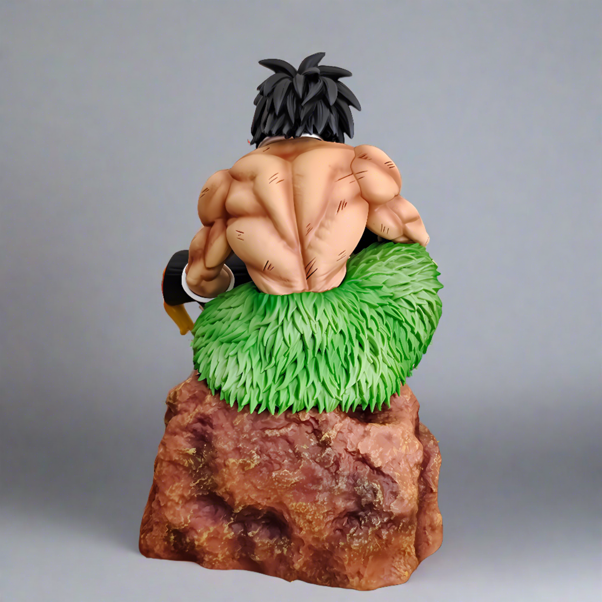 Rear view of the Dragon Ball collectible figure Broly seated on a rock, highlighting his muscular back and green fur waistband, set against a backdrop with sparks and embers.